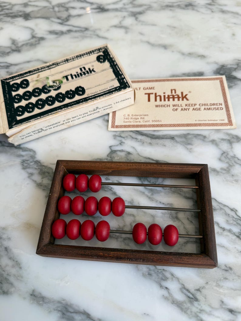 K&ouml;nnte beinhalten: Ein Vintage-Thimk-Spiel mit einem h&ouml;lzernen Abakus und einer Schachtel mit schwarzen Spielsteinen. Der Abakus hat einen dunklen Holzrahmen und rote Perlen. Die Spielbox und die Karte sind beige mit schwarzem und rotem Text.