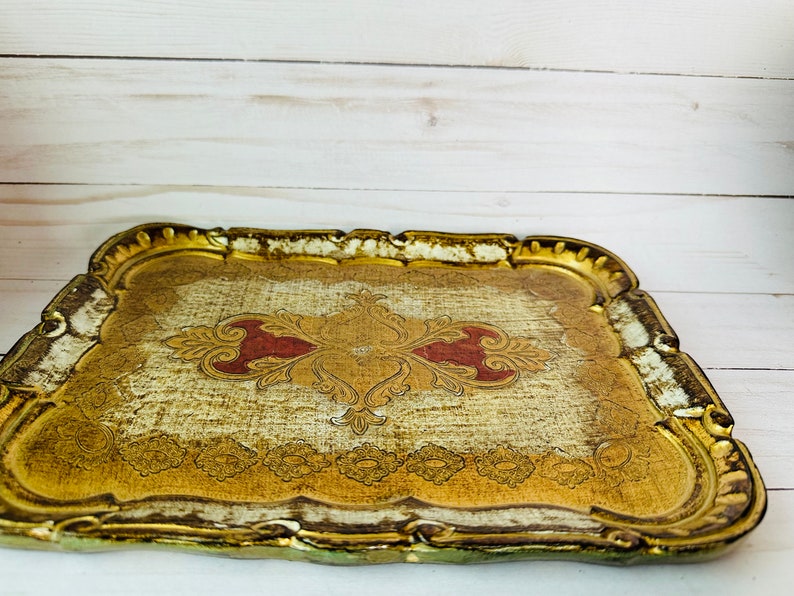 May include: A gold and white wooden tray with a decorative red and gold design. The tray has a scalloped edge and is perfect for serving snacks or drinks.