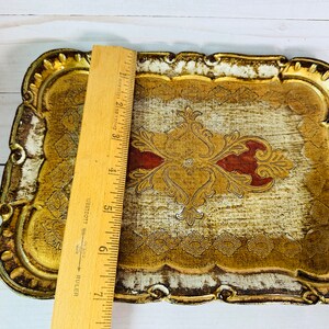 May include: A gold and white wooden tray with an ornate design. The tray has a raised edge and a scalloped border. The design features a central motif with red accents.