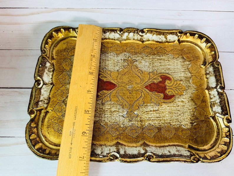 May include: A gold and white wooden tray with an ornate design. The tray has a raised edge and a scalloped border. The design features a central motif with red accents.
