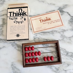 K&ouml;nnte beinhalten: Ein Vintage-Abakus aus Holz mit roten Perlen, zusammen mit einer Spielbox und einer Karte. Auf der Box steht "n Thimk THINK IT OVER!" und auf der Karte steht "AN ADULT GAME Thimk WHICH WILL KEEP CHILDREN OF ANY AGE AMUSED."