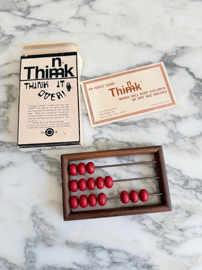 K&ouml;nnte beinhalten: Ein Vintage-Abakus aus Holz mit roten Perlen, zusammen mit einer Spielbox und einer Karte. Auf der Box steht "n Thimk THINK IT OVER!" und auf der Karte steht "AN ADULT GAME Thimk WHICH WILL KEEP CHILDREN OF ANY AGE AMUSED."
