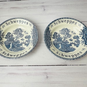 May include: Two vintage blue and white porcelain plates with a scenic design and the word "Schweppes" in a curved font. The plates feature a detailed landscape with pagodas and bridges.