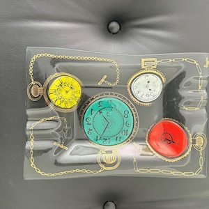 May include: Rectangular glass ashtray with four colorful pocket watch designs. The watches feature yellow, white, teal, and red faces with gold-colored chain details. The ashtray is clear with a slightly curved shape.