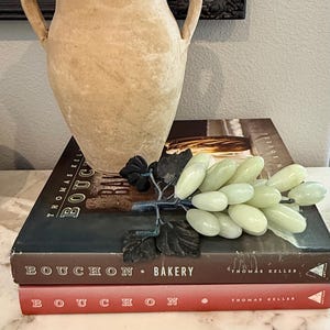 May include: A beige, two-handled vase sits atop two books, one red and one dark brown, with the title "Bouchon Bakery". A decorative bunch of light green grapes rests on the books, adding a touch of elegance to the composition.