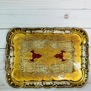 May include: A gold and white rectangular serving tray with an ornate design. The tray has a raised edge and a scalloped border. The design features a central motif of a stylized flower with red accents.