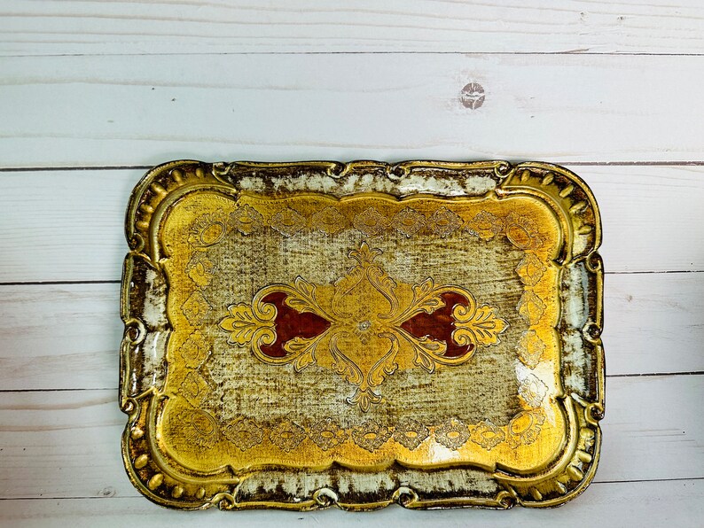 May include: A gold and white rectangular serving tray with an ornate design. The tray has a raised edge and a scalloped border. The design features a central motif of a stylized flower with red accents.