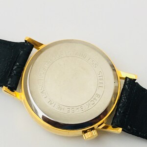 May include: A gold-toned wristwatch with a black leather band. The watch face is not visible, but the back is visible, with the words "Stainless Steel Back Base Metal Case Dustproof" printed in a circular pattern.