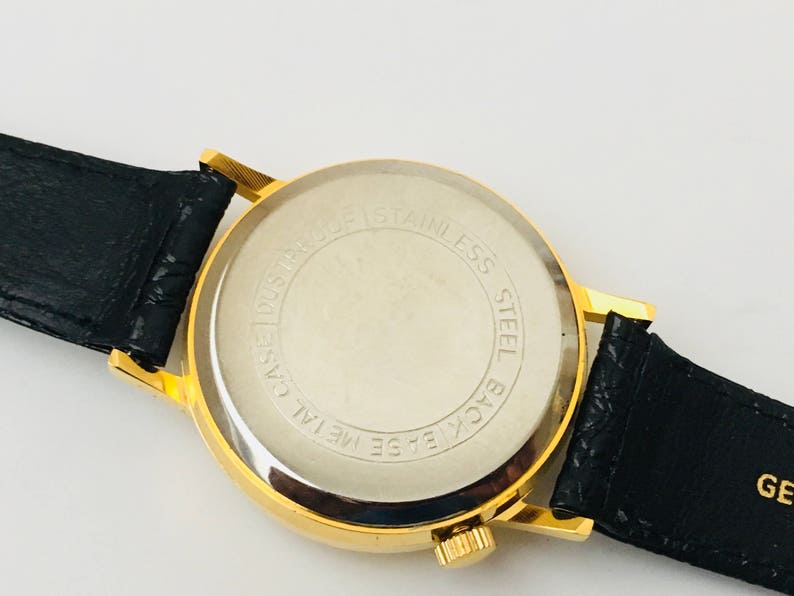 May include: A gold-toned wristwatch with a black leather band. The watch face is not visible, but the back is visible, with the words "Stainless Steel Back Base Metal Case Dustproof" printed in a circular pattern.