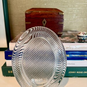 May include: Clear glass ashtray with a wavy, striped design. The oval-shaped ashtray has a small, circular base and a textured interior. The ashtray is sitting on a stack of books.