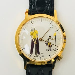 May include: Gold-tone wristwatch with a black leather band. The watch face features a golf-themed illustration with two figures, one swinging a club and the other holding a flag. The watch has a gold bezel.