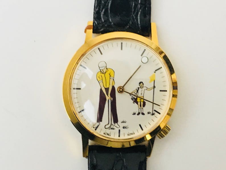 May include: Gold-tone wristwatch with a black leather band. The watch face features a golf-themed illustration with two figures, one swinging a club and the other holding a flag. The watch has a gold bezel.