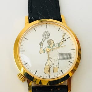 May include: Gold-tone wristwatch with a black leather band. The white watch face features a tennis player illustration with a racket and ball. The watch has a round face and a textured winding crown.