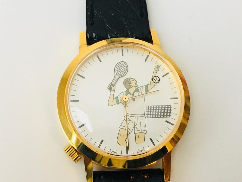 May include: Gold-tone wristwatch with a black leather band. The white watch face features a tennis player illustration with a racket and ball. The watch has a round face and a textured winding crown.
