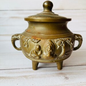 May include: A small, antique brass incense burner with a lid and three legs. The burner is decorated with intricate carvings of figures and trees.