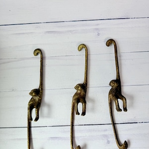 May include: Three gold monkey hooks hanging on a white wooden wall. Each hook is shaped like a monkey hanging upside down with its tail curled into a hook.