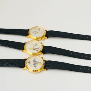 May include: Three gold-toned wristwatches with black leather bands are displayed. Each watch has a white face with a unique cartoon character design. The watches are arranged horizontally against a white background.