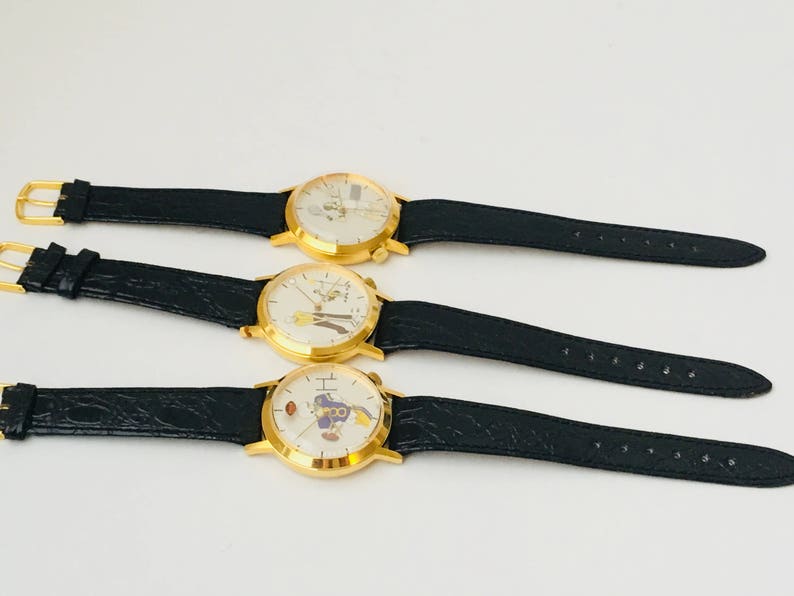 May include: Three gold-toned wristwatches with black leather bands are displayed. Each watch has a white face with a unique cartoon character design. The watches are arranged horizontally against a white background.