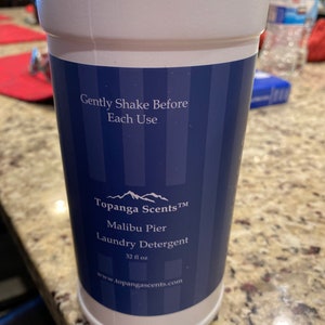 May include: A white plastic bottle of laundry detergent with a blue and white label. The label says "Topanga Scents TM Malibu Pier Laundry Detergent 32 fl oz www.topangascents.com".