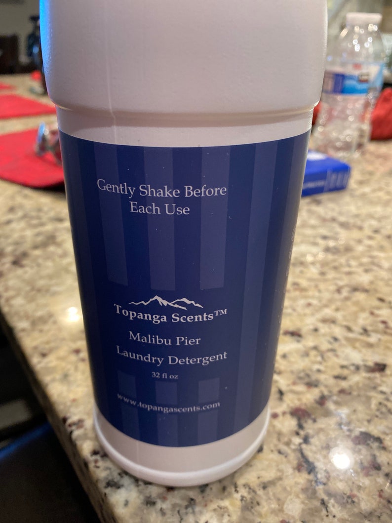 May include: A white plastic bottle of laundry detergent with a blue and white label. The label says "Topanga Scents TM Malibu Pier Laundry Detergent 32 fl oz www.topangascents.com".