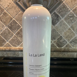 May include: A white plastic bottle of La La Land laundry detergent with a black cap. The bottle is labeled with the text "La La Land", "Laundry Detergent", "Powder | Peony | Linen", and "32 fl oz".