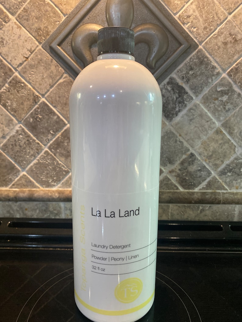 May include: A white plastic bottle of La La Land laundry detergent with a black cap. The bottle is labeled with the text "La La Land", "Laundry Detergent", "Powder | Peony | Linen", and "32 fl oz".