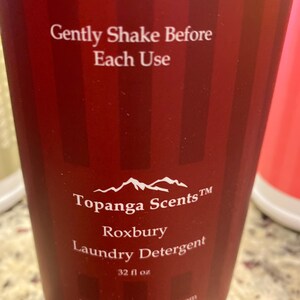 May include: A red and white striped bottle of laundry detergent with the text "Gently Shake Before Each Use" and "Topanga Scents TM Roxbury Laundry Detergent 32 fl oz www.topangascents.com".