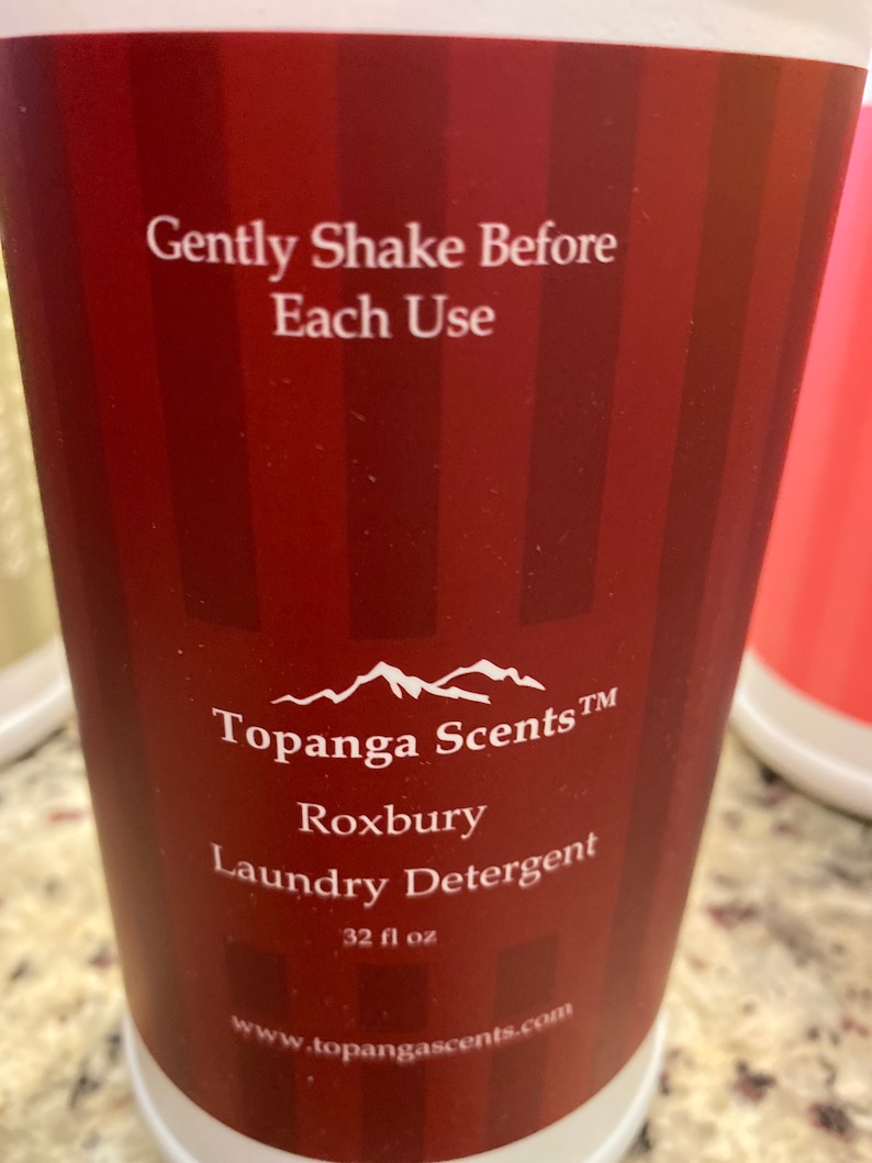May include: A red and white striped bottle of laundry detergent with the text "Gently Shake Before Each Use" and "Topanga Scents TM Roxbury Laundry Detergent 32 fl oz www.topangascents.com".