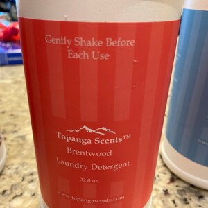 May include: A white plastic bottle with a red label. The label says "Topanga Scents TM Brentwood Laundry Detergent 32 fl oz www.topangascents.com". The label also has a mountain range graphic and the text "Gently Shake Before Each Use".