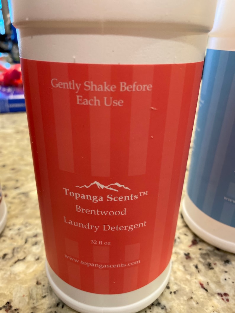 May include: A white plastic bottle with a red label. The label says "Topanga Scents TM Brentwood Laundry Detergent 32 fl oz www.topangascents.com". The label also has a mountain range graphic and the text "Gently Shake Before Each Use".