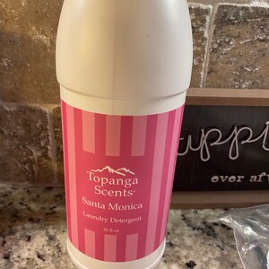 May include: A white plastic bottle with a pink and white striped label. The label says "Topanga Scents" and "Santa Monica Laundry Detergent" with a mountain range graphic. The bottle contains 32 fl oz.