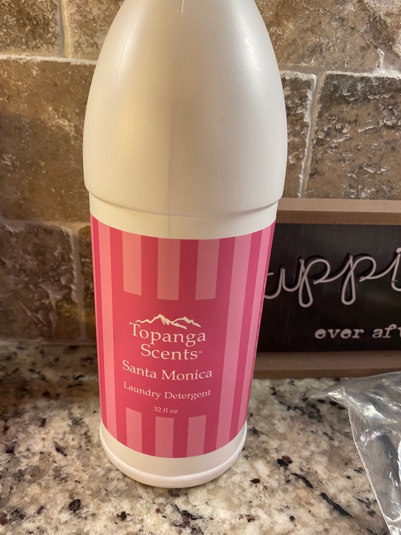 May include: A white plastic bottle with a pink and white striped label. The label says "Topanga Scents" and "Santa Monica Laundry Detergent" with a mountain range graphic. The bottle contains 32 fl oz.