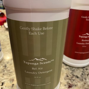 May include: White plastic bottle with a green and white striped label. The label reads "Topanga Scents&trade; Bel Air Laundry Detergent 32 fl oz www.topangascents.com". The bottle has the text "Gently Shake Before Each Use" on the label.