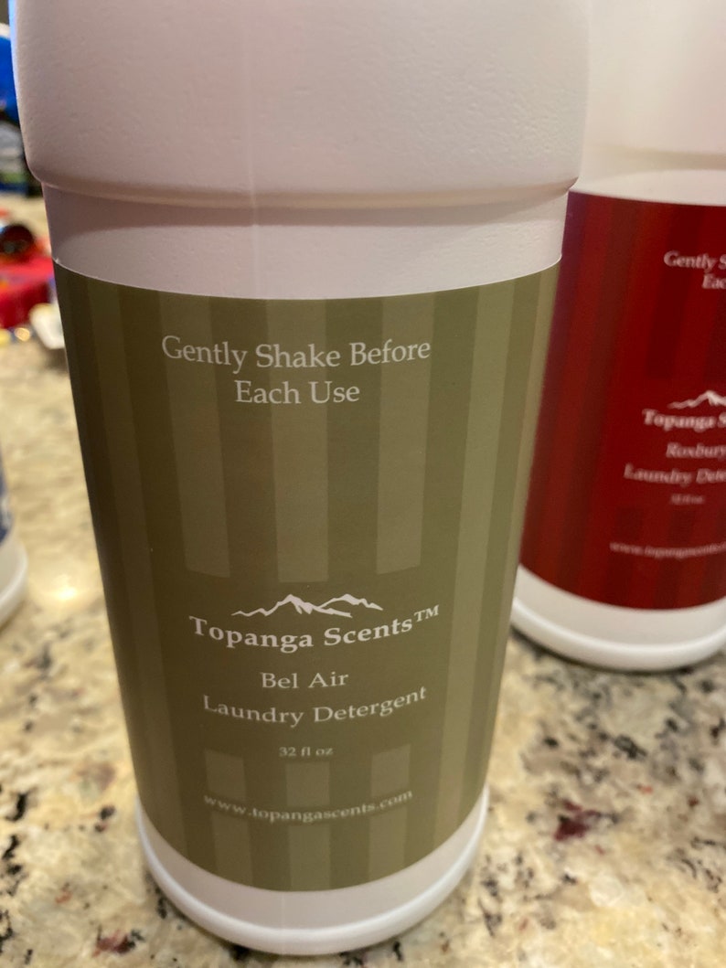 May include: White plastic bottle with a green and white striped label. The label reads "Topanga Scents&trade; Bel Air Laundry Detergent 32 fl oz www.topangascents.com". The bottle has the text "Gently Shake Before Each Use" on the label.