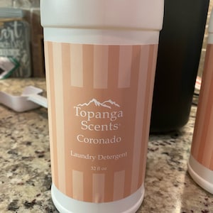 May include: A white plastic bottle with a pink and white striped label. The label says "Topanga Scents Coronado Laundry Detergent 32 fl oz."