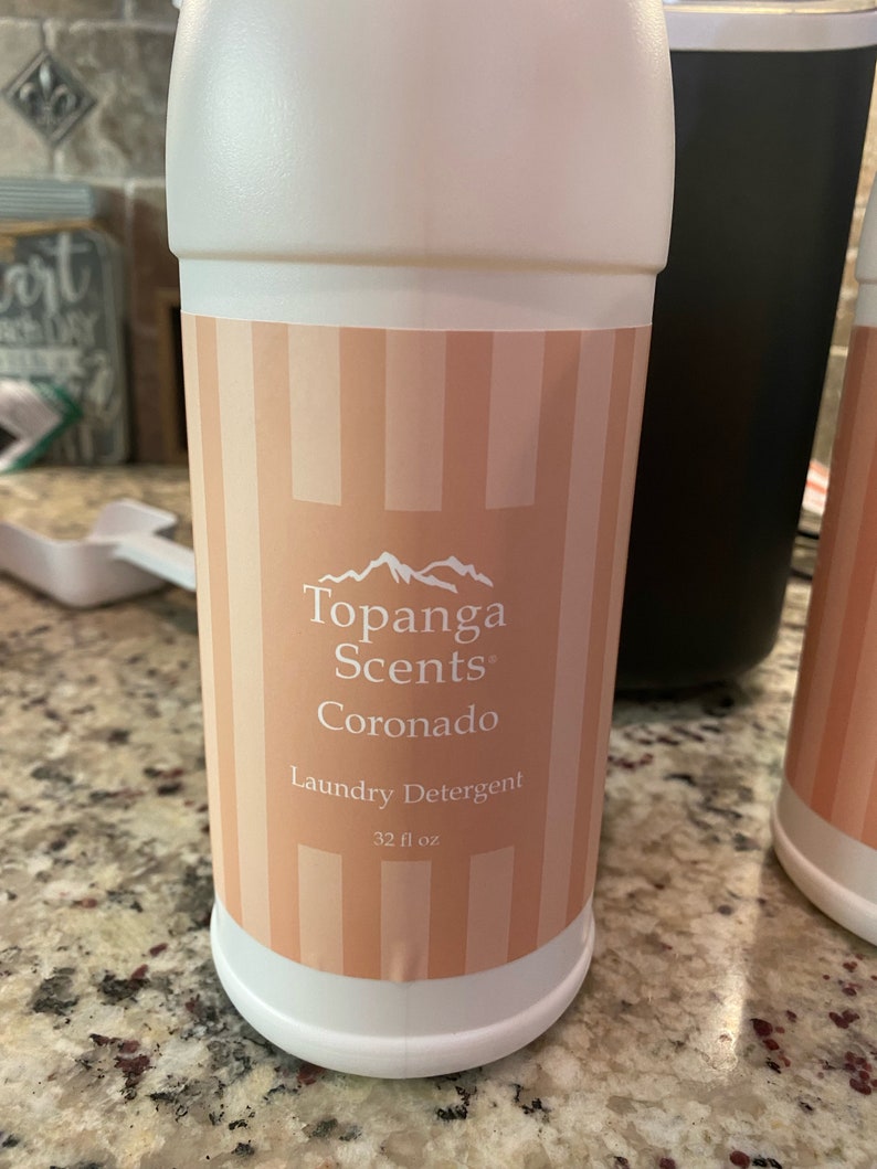 May include: A white plastic bottle with a pink and white striped label. The label says "Topanga Scents Coronado Laundry Detergent 32 fl oz."