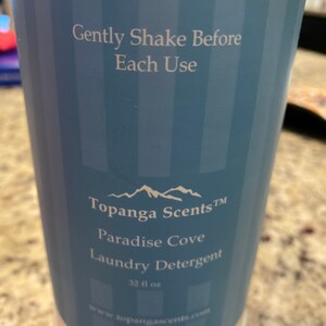 May include: A white plastic bottle of Topanga Scents Paradise Cove Laundry Detergent. The bottle is 32 fluid ounces and has a blue label with a mountain range logo. The label also says "Gently Shake Before Each Use" and "www.topangascents.com".