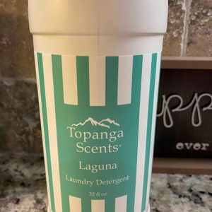 May include: White plastic bottle with a green and white striped label. The label reads "Topanga Scents" and "Laguna Laundry Detergent" with a mountain range logo. The bottle contains 32 fl oz.