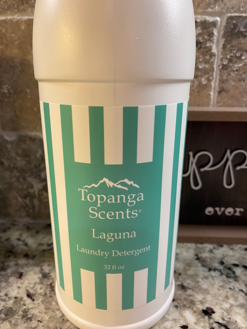 May include: White plastic bottle with a green and white striped label. The label reads "Topanga Scents" and "Laguna Laundry Detergent" with a mountain range logo. The bottle contains 32 fl oz.