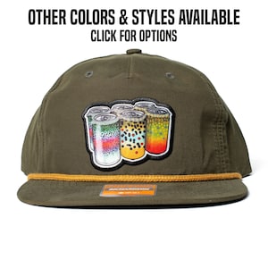 May include: An olive green baseball cap with a flat peak and a gold rope detail. The cap has a patch with colourful illustrations of beverage cans. The text "OTHER COLORS & STYLES AVAILABLE CLICK FOR OPTIONS" is printed above the cap.