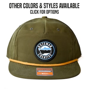 May include: A green baseball cap with a brown rope trim and a black and blue patch that says "Bozeman Creative" with a stylized outline of Montana. The patch is centered on the front of the cap.