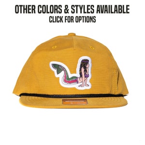 Könnte beinhalten: Eine Baseballkappe in Senfgelb mit schwarzem Schirm und einem Aufnäher einer Meerjungfrau mit grünem Schwanz und lila Haaren. Der Aufnäher trägt unten den Schriftzug "Richardson".