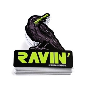 May include: A stack of black and green stickers with a raven holding a green stick in its beak. The sticker says "Ravin' by Bozeman Creative."