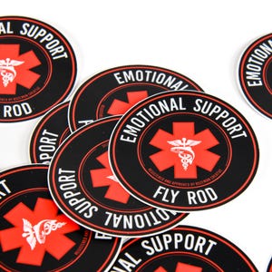 Może przedstawiać: Kolekcja czarnych i czerwonych okrągłych naklejek z białymi napisami "EMOTIONAL SUPPORT" i "FLY ROD". Naklejki mają czerwony symbol krzyża z białym kaduceuszem pośrodku. Naklejki są rozrzucone na białej powierzchni.