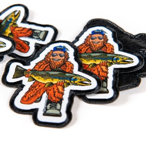 May include: A set of four iron-on patches featuring a cartoon sasquatch wearing a blue baseball cap and sunglasses, holding a large trout. The patches are white with black outlines and are designed to be sewn or ironed onto clothing or bags.
