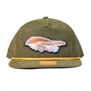 Puede incluir: Gorra de béisbol verde oliva con visera plana y detalle de cuerda amarilla. La gorra presenta un parche con una ilustración detallada de una mosca de pesca en tonos naranja, marrón y blanco. El nombre de la marca es visible en la visera.