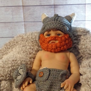 May include: A baby doll wearing a gray crocheted viking hat with orange beard, a gray crocheted diaper, and brown crocheted booties.