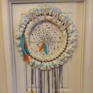 May include: A blue and white diaper wreath with the name "RYAN" spelled out in brown letters. The wreath is decorated with feathers and beads and hangs on a white door.