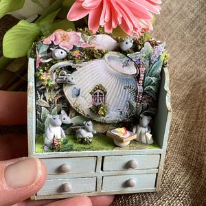 Puede incluir: Una casa de hadas en miniatura con una unidad de cajones azul claro. La casa presenta un diseño caprichoso con una estructura en forma de tetera, pequeñas figuras de ratones y una mesa con una rebanada de queso. Flores rosas y vegetación añaden un toque de color.