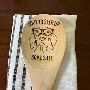 Könnte beinhalten: Ein Holzlöffel mit einem Hund, der eine Brille trägt, und dem Text "Bout to stir up some shit", der darauf graviert ist.
